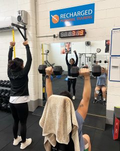 trainer and client looking in a mirror. client holds dumbells above their head.