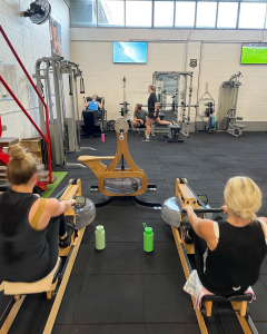 two women side by side on rowing machines while other people workout on gym equpiment in the background