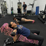 a woman using dumbells lying on a bench while another woman does a push-up