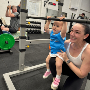 WOMAN HOLDING A YOUNG TODDLER UPRIGHT AT BARBELL while both looking in the mirror