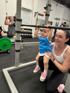 WOMAN HOLDING A YOUNG TODDLER UPRIGHT AT BARBELL while both looking in the mirror
