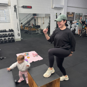 woman at a gym stepping onto a bench while a toddler plays happily next to her.