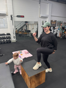 woman at a gym stepping onto a bench while a toddler plays happily next to her.