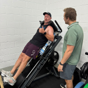 man on leg press machine doing squats and displaying thumbs up whilst personal trainer watches.